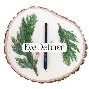 May include: A black eye definer makeup brush with a dark brown tip, featuring the text Moonrise Creek on the handle. The brush is centered between two green cedar sprigs on a light-colored circular wood slice with the text Eye Definer overlaid.
