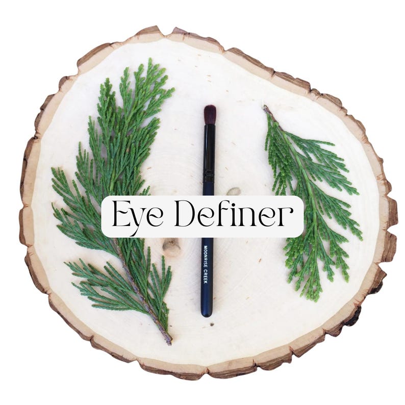May include: A black eye definer makeup brush with a dark brown tip, featuring the text Moonrise Creek on the handle. The brush is centered between two green cedar sprigs on a light-colored circular wood slice with the text Eye Definer overlaid.