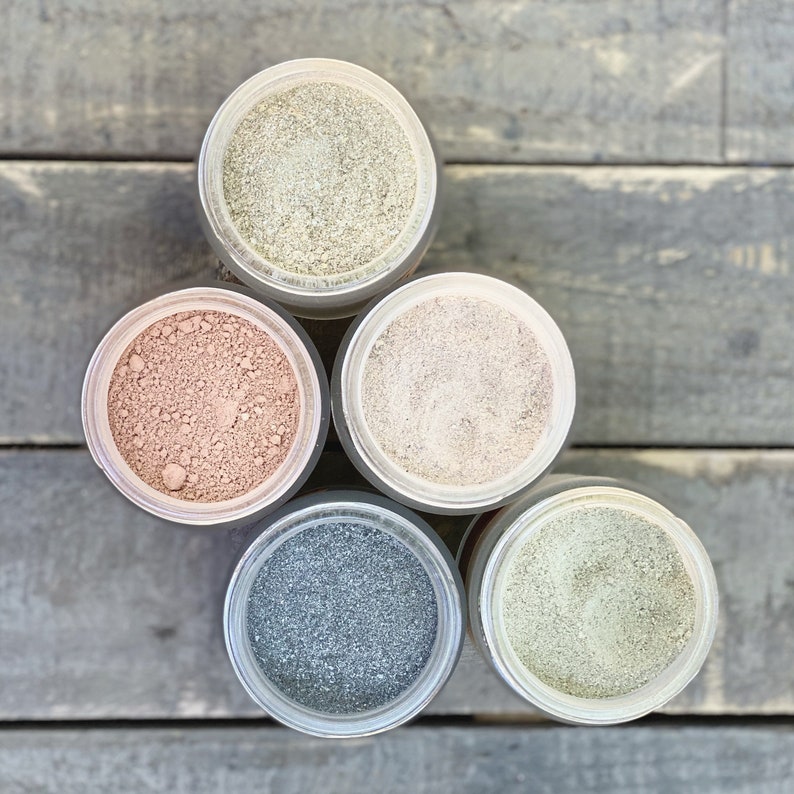 May include: Five glass jars with lids containing different colored powders. The jars are arranged in a triangle shape on a wooden surface. The powders are light brown, beige, gray, and green.