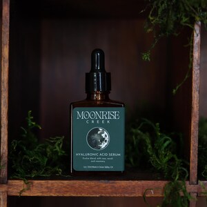 Op de afbeelding: Een bruine glazen fles Moonrise Creek Hyaluronzuur Serum met een zwarte druppelaar. Het etiket is teal met witte tekst en een maangrafiek. De fles staat in een houten plank met groen mos.