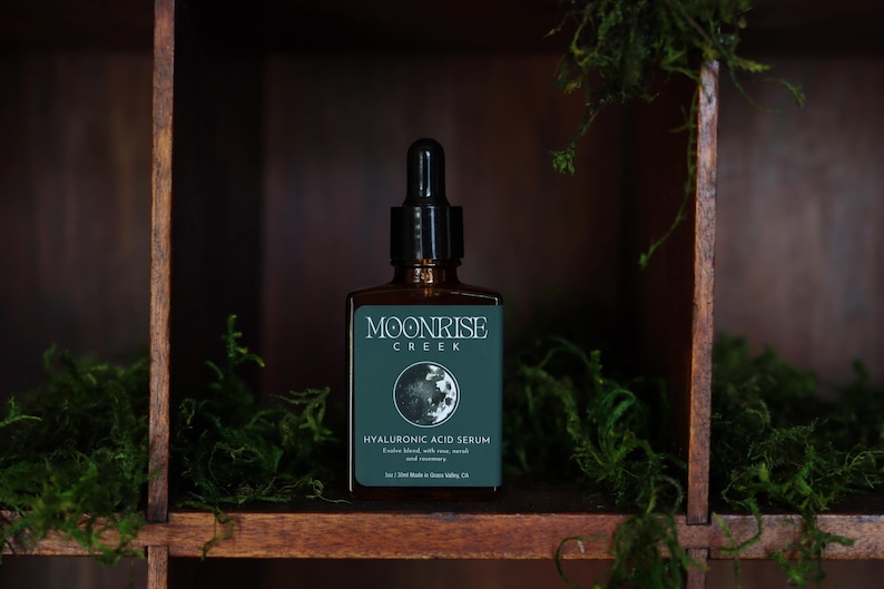 Op de afbeelding: Een bruine glazen fles Moonrise Creek Hyaluronzuur Serum met een zwarte druppelaar. Het etiket is teal met witte tekst en een maangrafiek. De fles staat in een houten plank met groen mos.