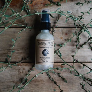 Könnte beinhalten: Eine klare Glasflasche mit schwarzem Pumpspender und einem braunen Etikett mit der Aufschrift "Moonrise Creek Herbal Apothecary & Botanical Skin Care" und "Luminous Body Oil". Das Etikett enthält auch den Text "Moonstone Sage, Sandalwood & Ylang Ylang". Die Flasche steht auf einer Holzoberfläche mit grünen, belaubten Zweigen.