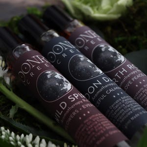 May include: Three aromatherapy roll-on bottles with black caps and labels featuring a moon design. The bottles are labeled "Wild Spirit", "Moon Dearth", and "Earth Rose". The bottles are surrounded by greenery and white flowers.