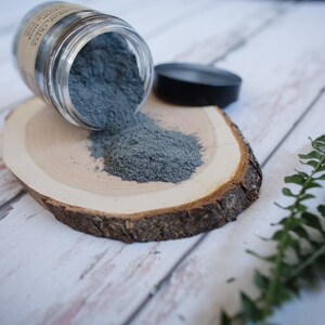 May include: A glass jar with a gray powder spilling out onto a wooden slice. The powder is a light gray color and is spread out on the wood. There is a green fern in the background.