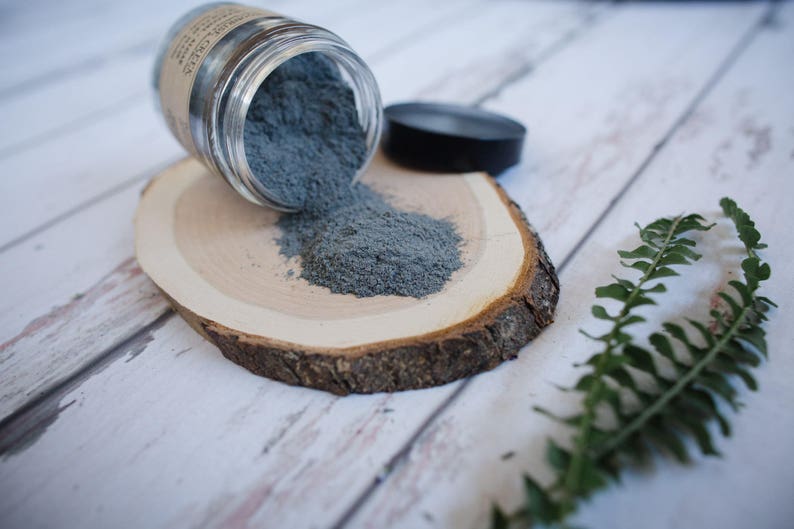 May include: A glass jar with a gray powder spilling out onto a wooden slice. The powder is a light gray color and is spread out on the wood. There is a green fern in the background.