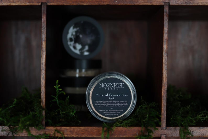 May include: A jar of Moonrise Creek Mineral Foundation in the shade "Fair" is displayed on a wooden shelf. The jar has a black lid with white text. Other jars are visible in the background, along with green moss.