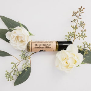 Puede incluir: Una pequeña botella de vidrio marrón con un aplicador de bola negra. La botella está etiquetada como "Tension Ease Aromatherapy Oil Blend". La botella está rodeada de rosas blancas y hojas verdes de eucalipto.