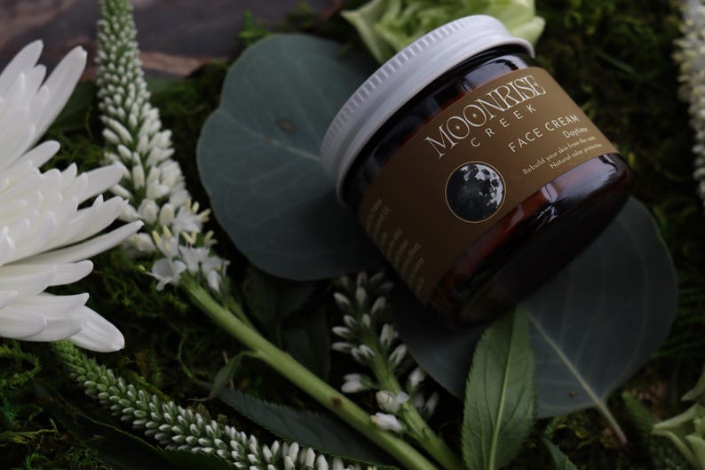 Puede incluir: Un tarro de crema facial Moonrise Creek con tapa blanca y etiqueta marr&oacute;n con un gr&aacute;fico de luna. El tarro est&aacute; rodeado de flores blancas, hojas verdes y musgo. El texto de la etiqueta dice "Face Cream Daytime".