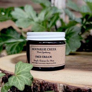 May include: A small, dark glass jar with a white lid, labeled "MOONRISE CREEK FACE CREAM." The jar sits on a wooden surface, surrounded by green foliage. The label also reads "Simple Skincare for Men" and "Non Pore Clogging Formulation."