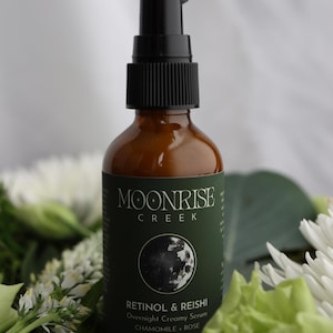 K&ouml;nnte beinhalten: Eine braune Glasflasche mit einem schwarzen Pumpspender. Die Flasche hat ein gr&uuml;nes Etikett mit dem Text "Moonrise Creek" und "Retinol & Reishi Overnight Creamy Serum". Die Flasche ist von wei&szlig;en und gr&uuml;nen Blumen umgeben.