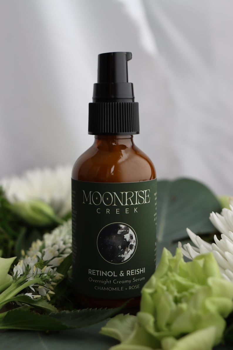K&ouml;nnte beinhalten: Eine braune Glasflasche mit einem schwarzen Pumpspender. Die Flasche hat ein gr&uuml;nes Etikett mit dem Text "Moonrise Creek" und "Retinol & Reishi Overnight Creamy Serum". Die Flasche ist von wei&szlig;en und gr&uuml;nen Blumen umgeben.