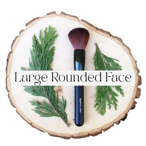 May include: A makeup brush with a black handle and dark brown bristles, featuring the text "Moonrise Creek" on the handle and the text "Large Rounded Face" overlaid across the center of the image, flanked by two green evergreen sprigs.