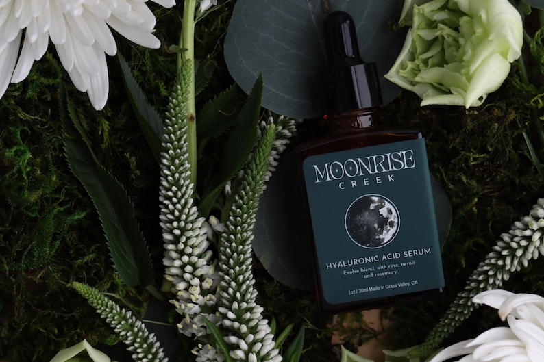 Op de afbeelding: Een bruine glazen fles Moonrise Creek Hyaluronzuur Serum met een zwarte druppelaar, geplaatst tegen een achtergrond van witte bloemen, groen gebladerte en mos. Het teal label toont een maan afbeelding en productdetails.