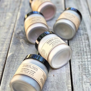 May include: Five glass jars with black lids containing different types of clay face masks. The jars are labeled with the brand name "Moonrise Creek" and the type of clay mask. The jars are arranged on a wooden surface.