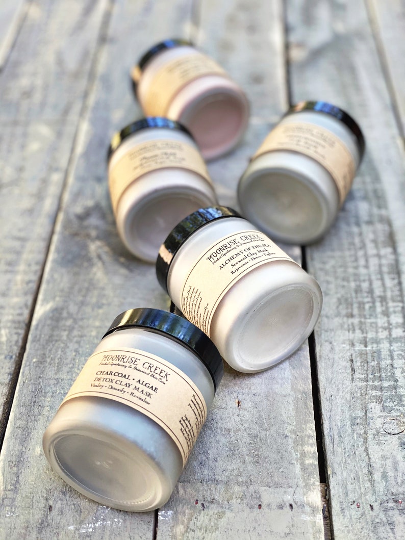 May include: Five glass jars with black lids containing different types of clay face masks. The jars are labeled with the brand name "Moonrise Creek" and the type of clay mask. The jars are arranged on a wooden surface.