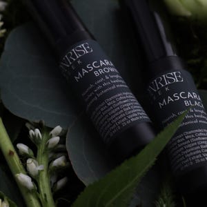 Puede incluir: Dos tubos de máscara de pestañas negros, etiquetados como "Sunrise Tek", con "Mascara Brown" y "Mascara Black". Los tubos están rodeados de vegetación y flores blancas. Cada tubo contiene 7,39 ml.