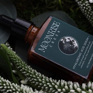 Op de afbeelding: Een bruine glazen fles met een zwarte druppelaar, met het label "MOONRISE CREEK HYALURONIC ACID SERUM". Het label bevat een maangrafiek en tekst over de ingredi&euml;nten van het product. De fles is omgeven door groen gebladerte en witte bloemen.