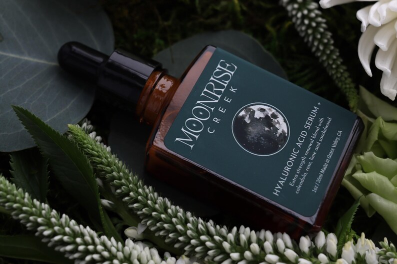 Op de afbeelding: Een bruine glazen fles met een zwarte druppelaar, met het label "MOONRISE CREEK HYALURONIC ACID SERUM". Het label bevat een maangrafiek en tekst over de ingredi&euml;nten van het product. De fles is omgeven door groen gebladerte en witte bloemen.