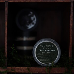 May include: A jar of Moonrise Creek translucent face powder is displayed in a dark wooden shelf compartment. The jar has a black lid and a label with white text. Green moss and small plants are in the foreground. Another jar is visible in the background.
