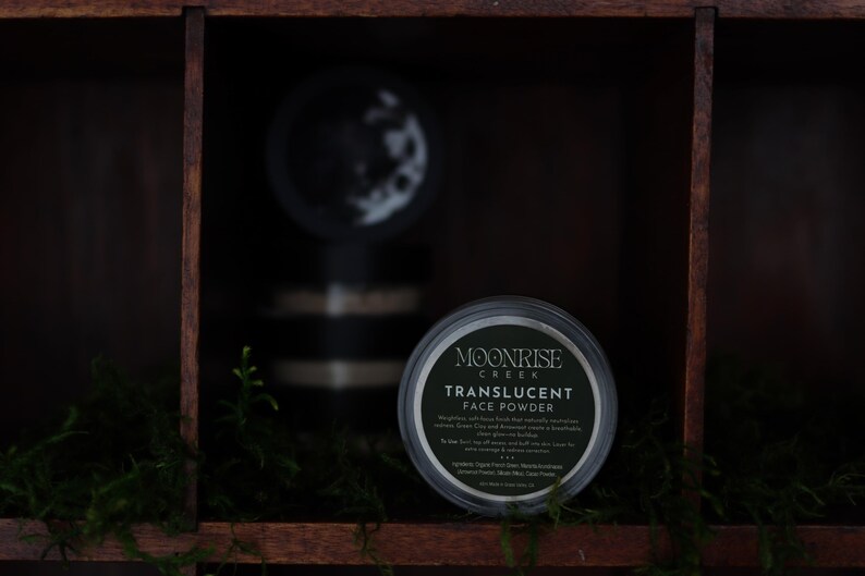 May include: A jar of Moonrise Creek translucent face powder is displayed in a dark wooden shelf compartment. The jar has a black lid and a label with white text. Green moss and small plants are in the foreground. Another jar is visible in the background.