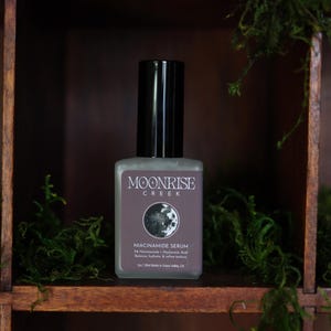Op de afbeelding: Een vierkante, matglazen fles Moonrise Creek Niacinamide Serum met een zwarte dop. Het etiket toont de merknaam, een maangrafiek en productdetails. De fles staat tegen een donkere houten achtergrond met groen mos.
