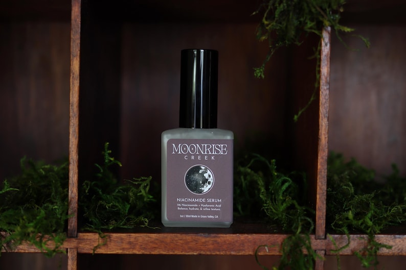 May include: A square, frosted glass bottle of Moonrise Creek Niacinamide Serum with a black cap. The label features the brand name, a moon graphic, and product details. The bottle is set against a dark wooden background with green moss.
