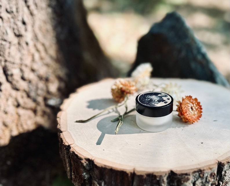 May include: A small, round jar with a black lid and a moon design on the lid. The jar is sitting on a wooden surface with dried flowers.