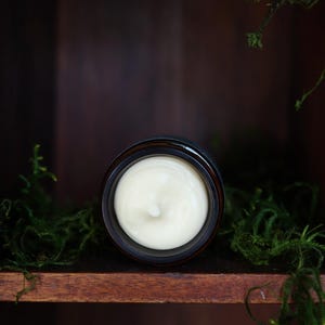 Puede incluir: Un tarro de crema de color beige claro, sobre un fondo de madera oscura y musgo verde. La crema est&aacute; en un tarro marr&oacute;n oscuro, evocando una est&eacute;tica natural y org&aacute;nica.