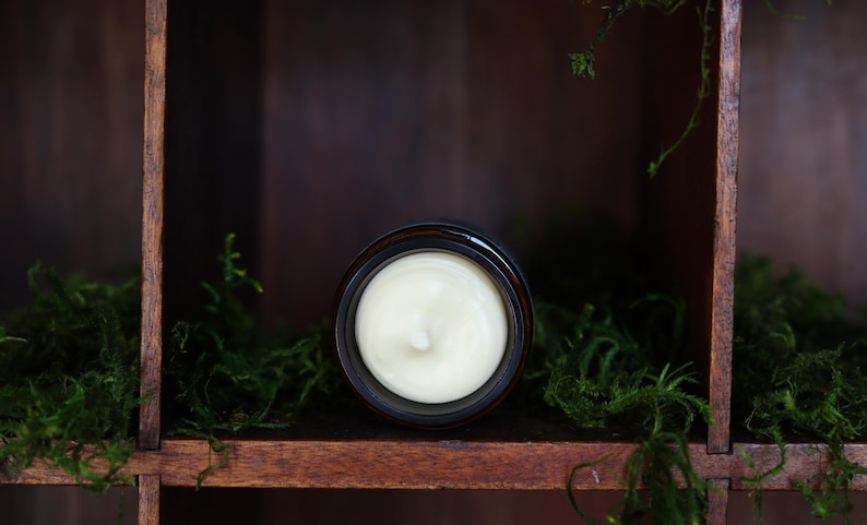 Puede incluir: Un tarro de crema de color beige claro, sobre un fondo de madera oscura y musgo verde. La crema est&aacute; en un tarro marr&oacute;n oscuro, evocando una est&eacute;tica natural y org&aacute;nica.