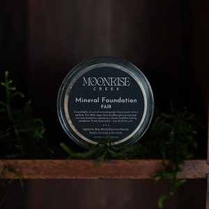 May include: A round container of Moonrise Creek Mineral Foundation in the shade "Fair." The container has a black label with white text. The container sits on a wooden shelf with green moss.