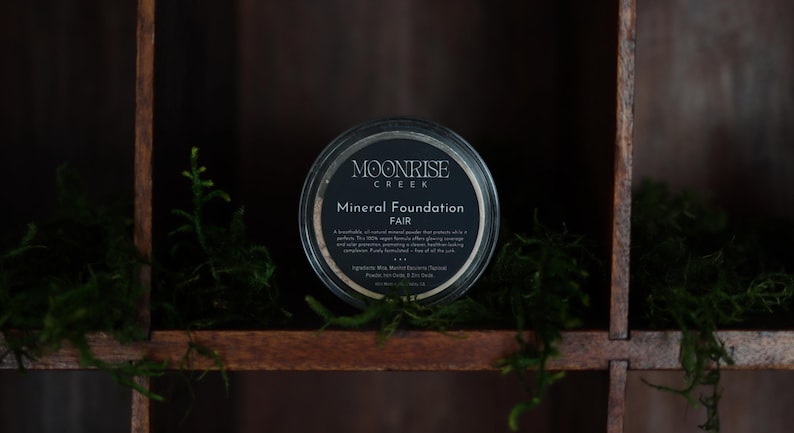 May include: A round container of Moonrise Creek Mineral Foundation in the shade "Fair." The container has a black label with white text. The container sits on a wooden shelf with green moss.