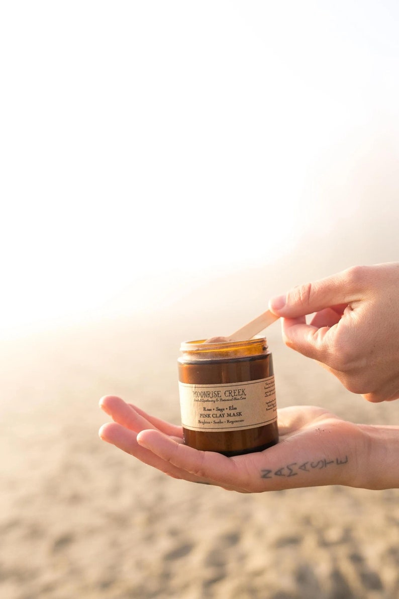 May include: A brown glass jar with a wooden spoon inside. The jar is labeled "Moonrise Creek" and "Pink Clay Mask". The jar is being held in a hand with a tattoo on the back.