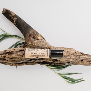 May include: A small brown glass bottle with a black rollerball top. The bottle is labeled "MOONRISE GLEE" and "MOON DANCE Aromatherapy Oil Blend". The bottle is sitting on a piece of driftwood with green leaves.
