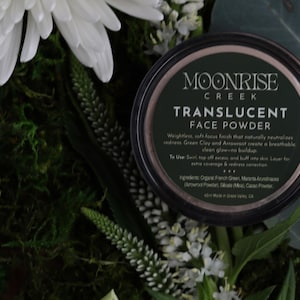 May include: A dark container of Moonrise Creek Translucent Face Powder is surrounded by white flowers, green foliage, and moss. The container's label displays the brand name and product details. The powder is designed to reduce redness and create a breathable glow.