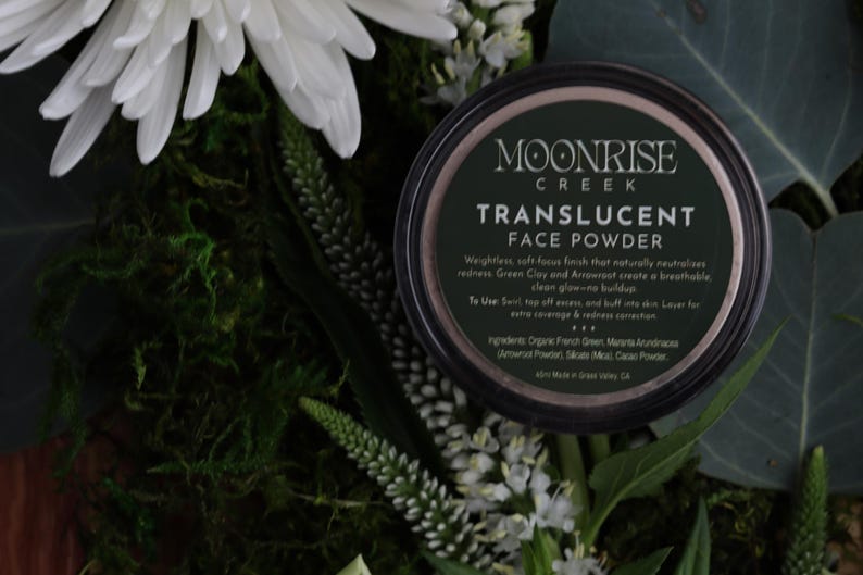 May include: A dark container of Moonrise Creek Translucent Face Powder is surrounded by white flowers, green foliage, and moss. The container's label displays the brand name and product details. The powder is designed to reduce redness and create a breathable glow.