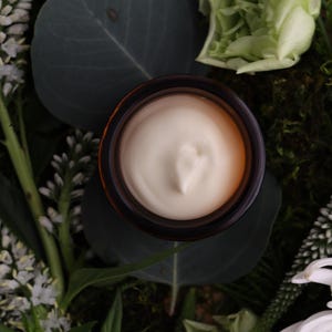 Puede incluir: Un tarro de crema se encuentra en medio de vegetaci&oacute;n y flores blancas. La crema es de color beige claro y el tarro tiene un borde marr&oacute;n oscuro. El arreglo incluye hojas verdes, musgo y flores blancas parecidas a margaritas.