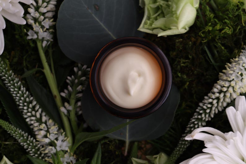 Puede incluir: Un tarro de crema se encuentra en medio de vegetaci&oacute;n y flores blancas. La crema es de color beige claro y el tarro tiene un borde marr&oacute;n oscuro. El arreglo incluye hojas verdes, musgo y flores blancas parecidas a margaritas.