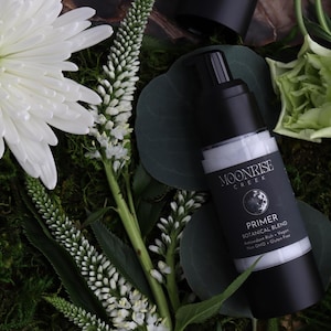 Könnte beinhalten: Eine schwarz-weiße Moonrise Creek Primer-Flasche mit einem Etikett für botanische Mischungen, umgeben von weißen Blumen und Grün. Auf dem Etikett steht "Primer Botanical Blend Antioxidant Rich + Vegan Non GMO + Gluten Free."