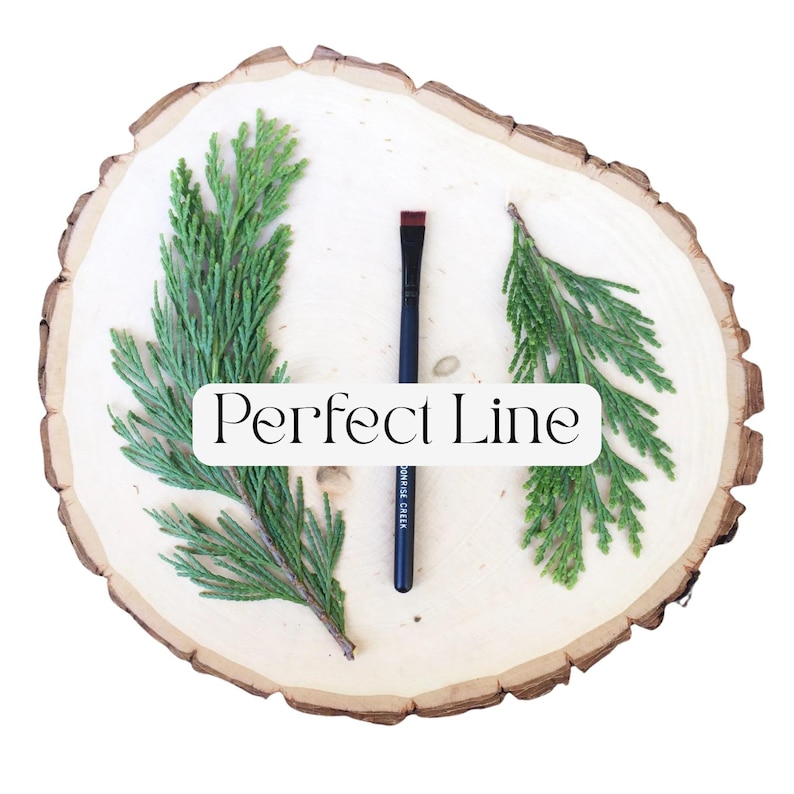 May include: A makeup brush with a black handle and a flat, angled red tip, positioned vertically between two green cedar sprigs on a light-colored wood slice. The text "Perfect Line" is printed in black on a white rectangular overlay.