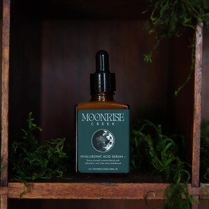 Op de afbeelding: Een bruine glazen fles Moonrise Creek Hyaluronzuur Serum met een zwarte druppelaar. Het etiket is teal met witte tekst en een maangrafiek. De fles staat op een houten plank met groen mos.