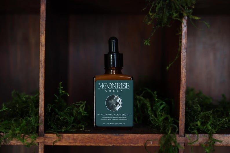 Op de afbeelding: Een bruine glazen fles Moonrise Creek Hyaluronzuur Serum met een zwarte druppelaar. Het etiket is teal met witte tekst en een maangrafiek. De fles staat op een houten plank met groen mos.