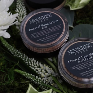 May include: Two round containers of Moonrise Creek Mineral Foundation, labelled "Fair" and "Matte Fair." The containers are clear with black labels and sit on greenery and white flowers. The text on the label is visible.