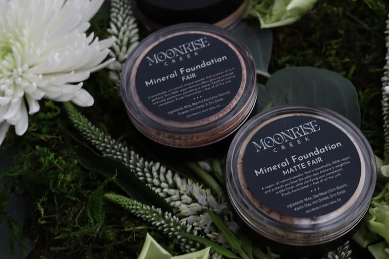 May include: Two round containers of Moonrise Creek Mineral Foundation, labelled "Fair" and "Matte Fair." The containers are clear with black labels and sit on greenery and white flowers. The text on the label is visible.