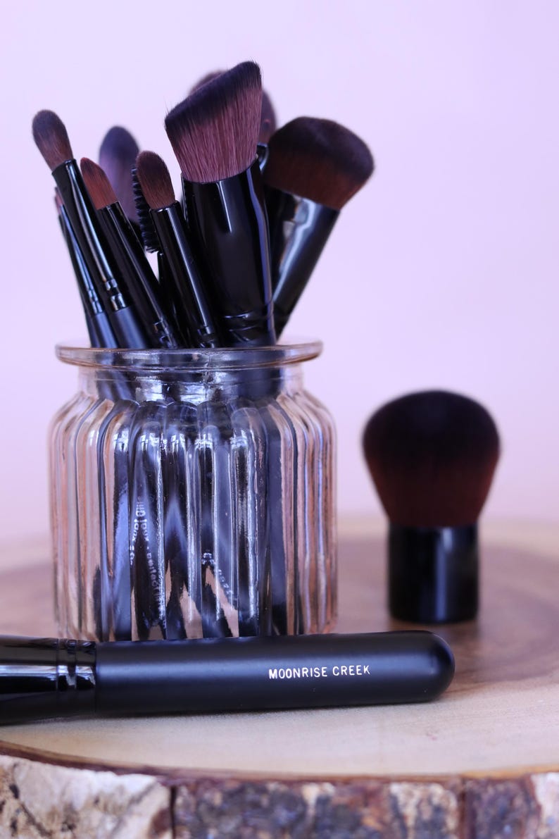 May include: A collection of makeup brushes with black handles and dark brown bristles, stored in a clear ribbed glass jar. One brush lies horizontally in the foreground with the text MOONRISE CREEK printed in white on its black handle.