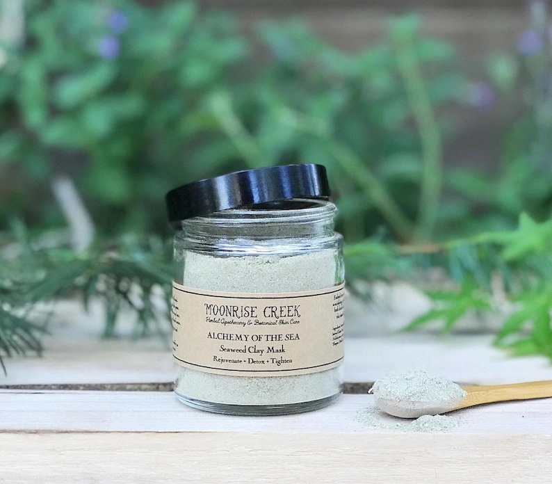 May include: A glass jar of green seaweed clay mask with a black lid. The label reads "Moonrise Creek Herbal Apothecary & Botanical Skin Care Alchemy of the Sea Seaweed Clay Mask Rejuvenate - Detox - Tighten"