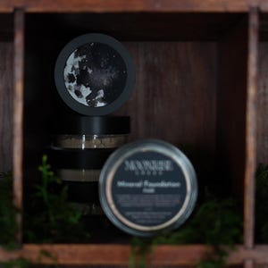 May include: A display of Moonrise Cosmetics mineral foundation products. The image features several black jars with labels, some stacked, and a circular lid with a moon design. The products are arranged in a wooden display case with green moss.