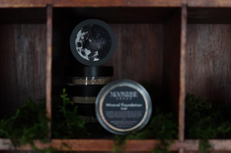 May include: A display of Moonrise Cosmetics mineral foundation products. The image features several black jars with labels, some stacked, and a circular lid with a moon design. The products are arranged in a wooden display case with green moss.