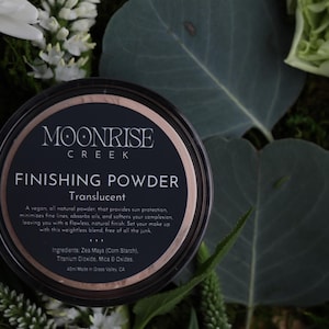May include: A black and rose gold finishing powder container with the text "MOONRISE CREEK FINISHING POWDER Translucent". The container is surrounded by greenery and white flowers. The product is a vegan, all-natural powder.