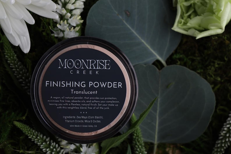 May include: A black and rose gold finishing powder container with the text "MOONRISE CREEK FINISHING POWDER Translucent". The container is surrounded by greenery and white flowers. The product is a vegan, all-natural powder.