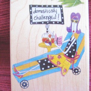 Op de afbeelding: Houten blok met een cartoon illustratie van een vrouw die ontspant op een ligstoel. Ze leest een boek met de tekst "Relax Your Broom" en heeft een drankje op een tafeltje. Het bord boven haar zegt "domestically challenged!"